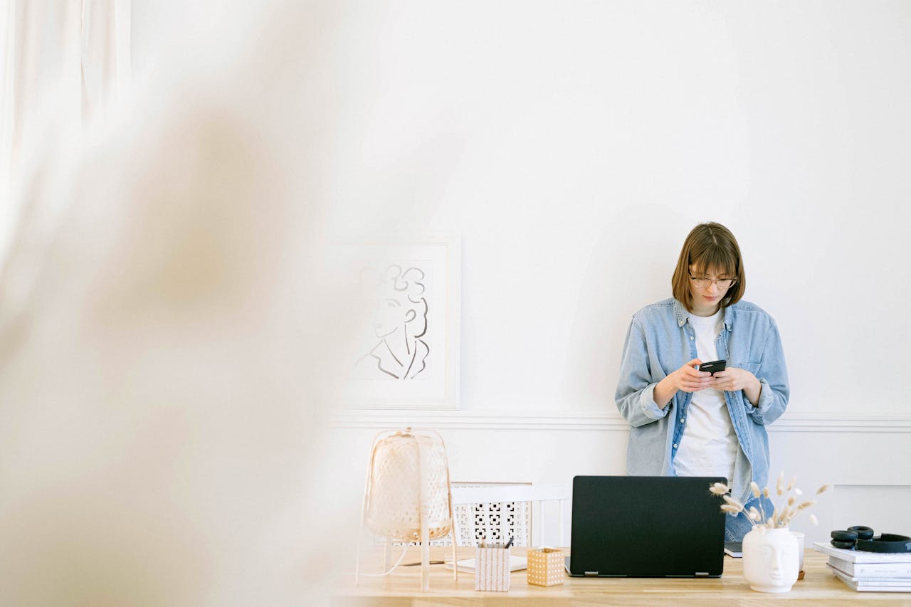 gallery-05 Woman in casual denim texting on smartphone in a bright, minimalist home office setup.